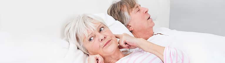 old woman plugging ears with an irritated expression laying in bed next to her partner